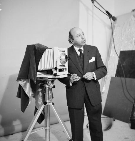 black and white image of Yousuf Karsh in a suit standing with an old fashioned camera.