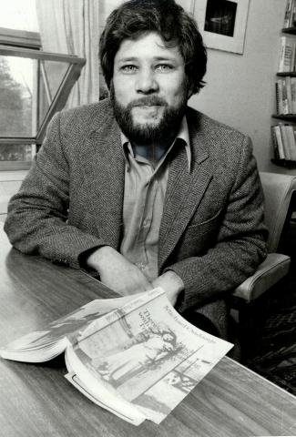 black and white image of Michael Ondaatje sitting at a table with an open book before him.