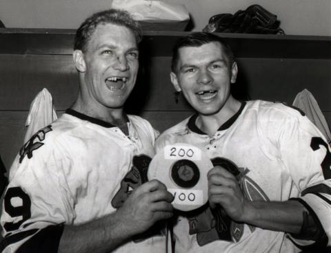Stan Mikita and another man in hockey jerseys with missing front teeth hold a puck between them.