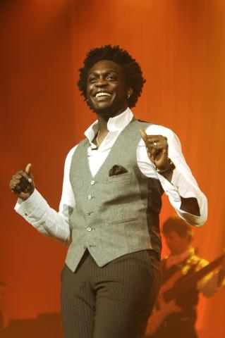 Cornelius Nyungura in a shirt and grey vest singing on stage .