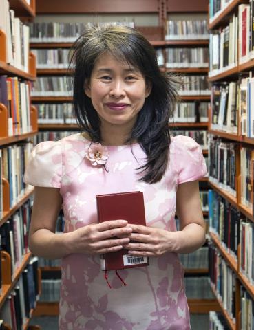 Kim Thúy wearing a pink dress stands among bookshelves holding a book.