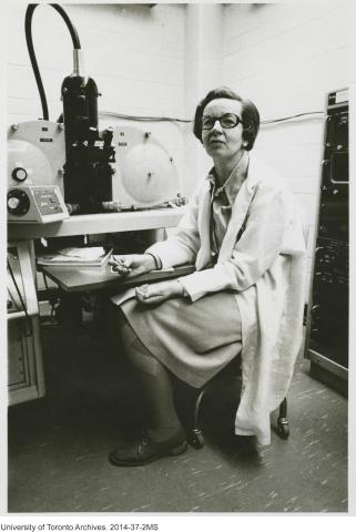 A black and white photo of a scientist in a lab coat, sitting in front of a large piece of lab equipment.