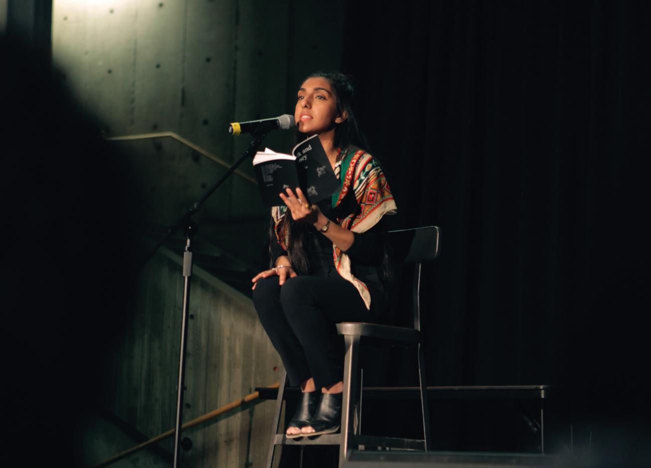 Rupi Kaur on stage seated on a high chair holding a book.