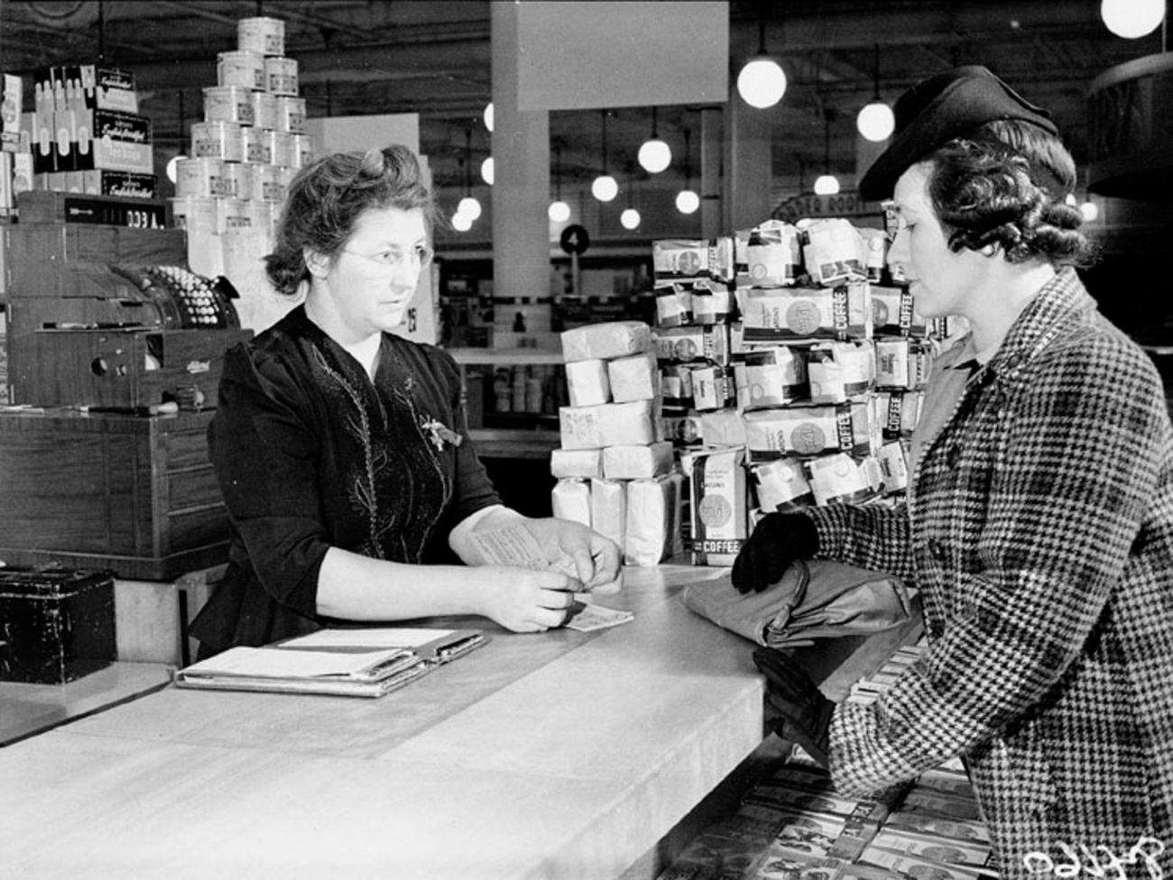 two women talk across a shop counter with piles of products behind them.