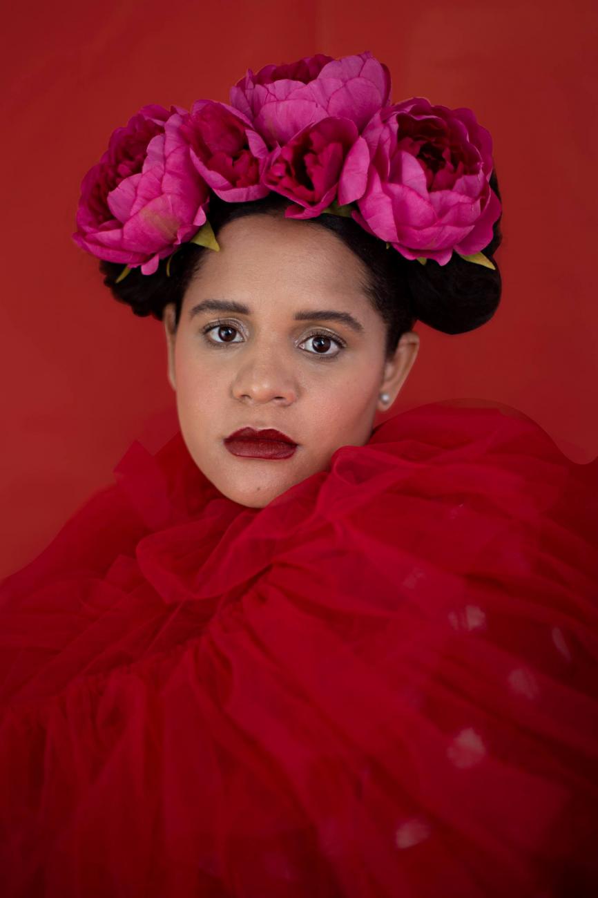 Head and shoulders photo of Lido Pimienta, her body covered in red netting, her hair up with large pink flowers in it.