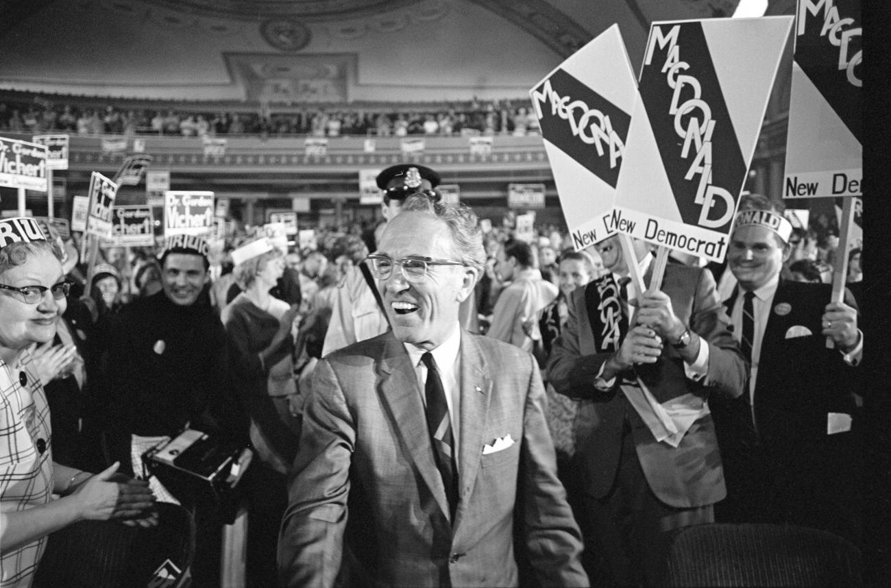 black and white photo of Tommy Douglas in a theatre with people behind him holding up signs.