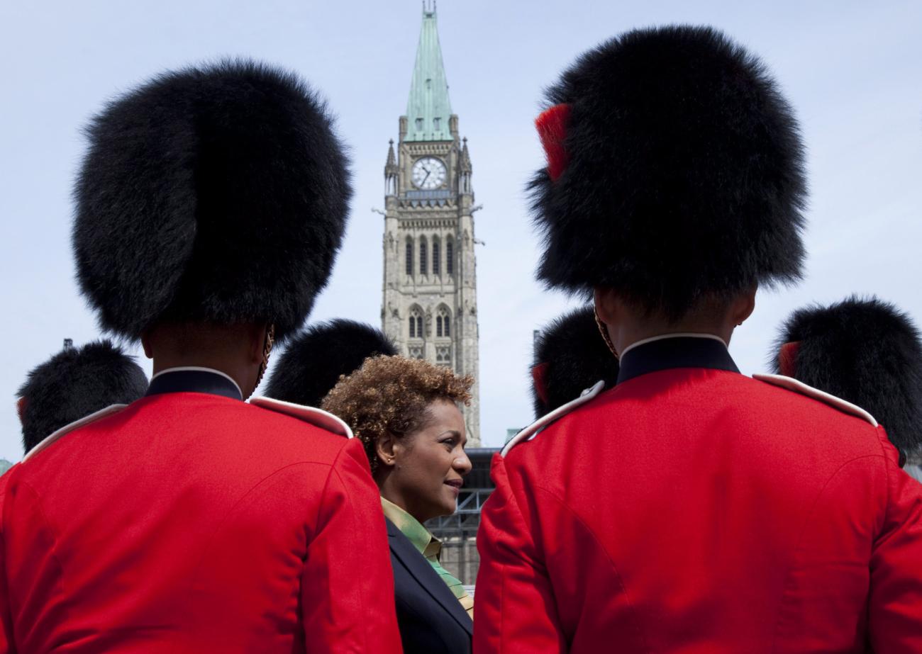 back view of two soldiers in red wearing bear-skin helmets, Michaëlle Jean and a tall clock tower can be seen between them.