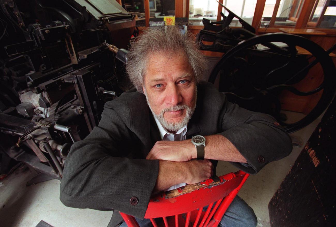 Michael Ondaatje sitting leaning on his arms on the back of a red chair.