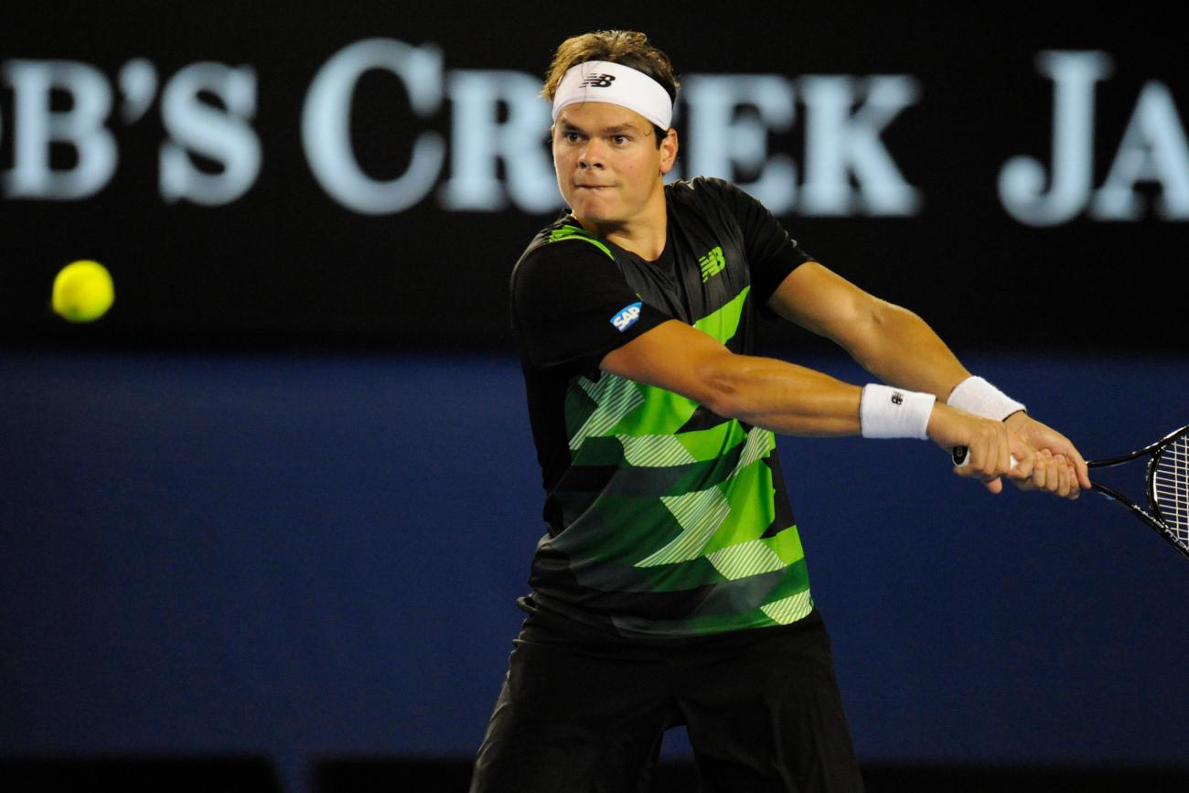 Milos Raonic wearing blue and green on tennis court swinging tennis racket at yellow ball.