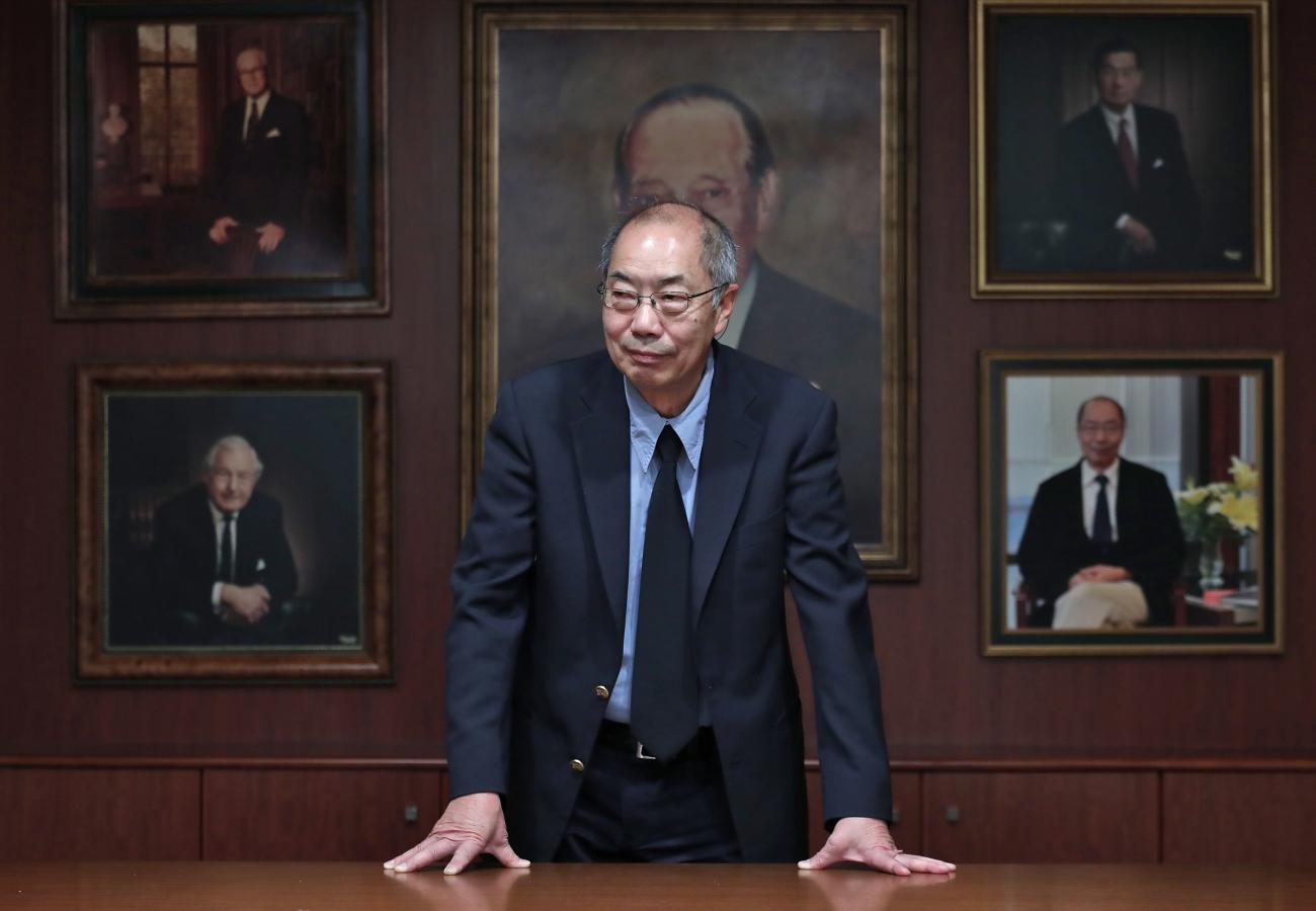Tak Wah Mak in dark suit leans on a desk with painted portraits of men on the wall behind him.