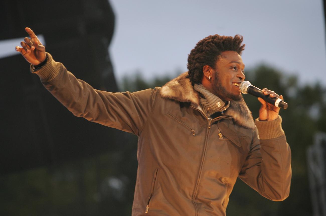 Corneille on stage outdoors singing with his arm extended, wearing a brown coat.