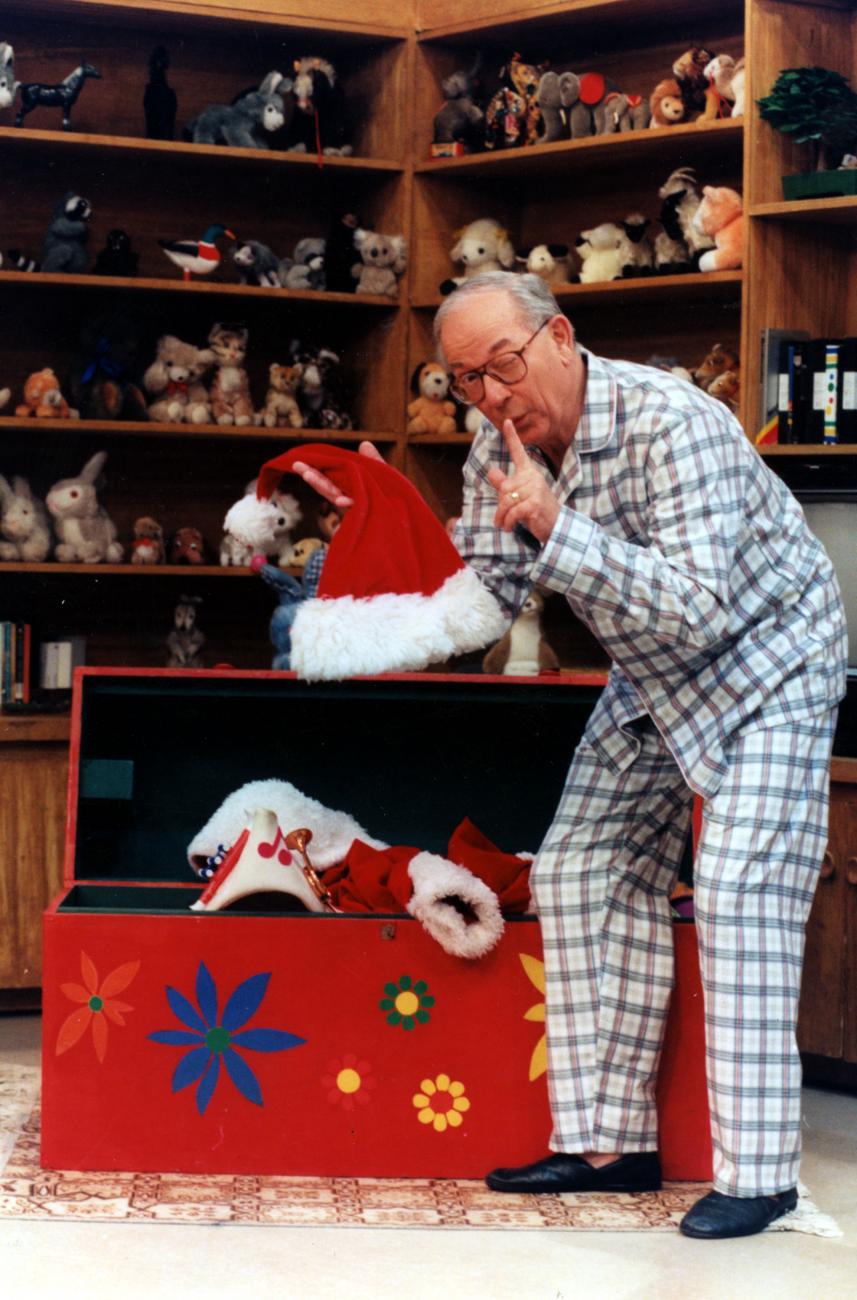 Ernie Coombs as Mr. Dressup in pyjamas pulling a Santa hat from a trunk in a room lined with shelves of toys.