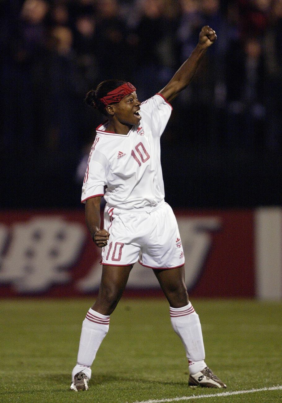 Charmaine Hooper in a white t-shirt and shorts on a soccer pitch with arm raised.