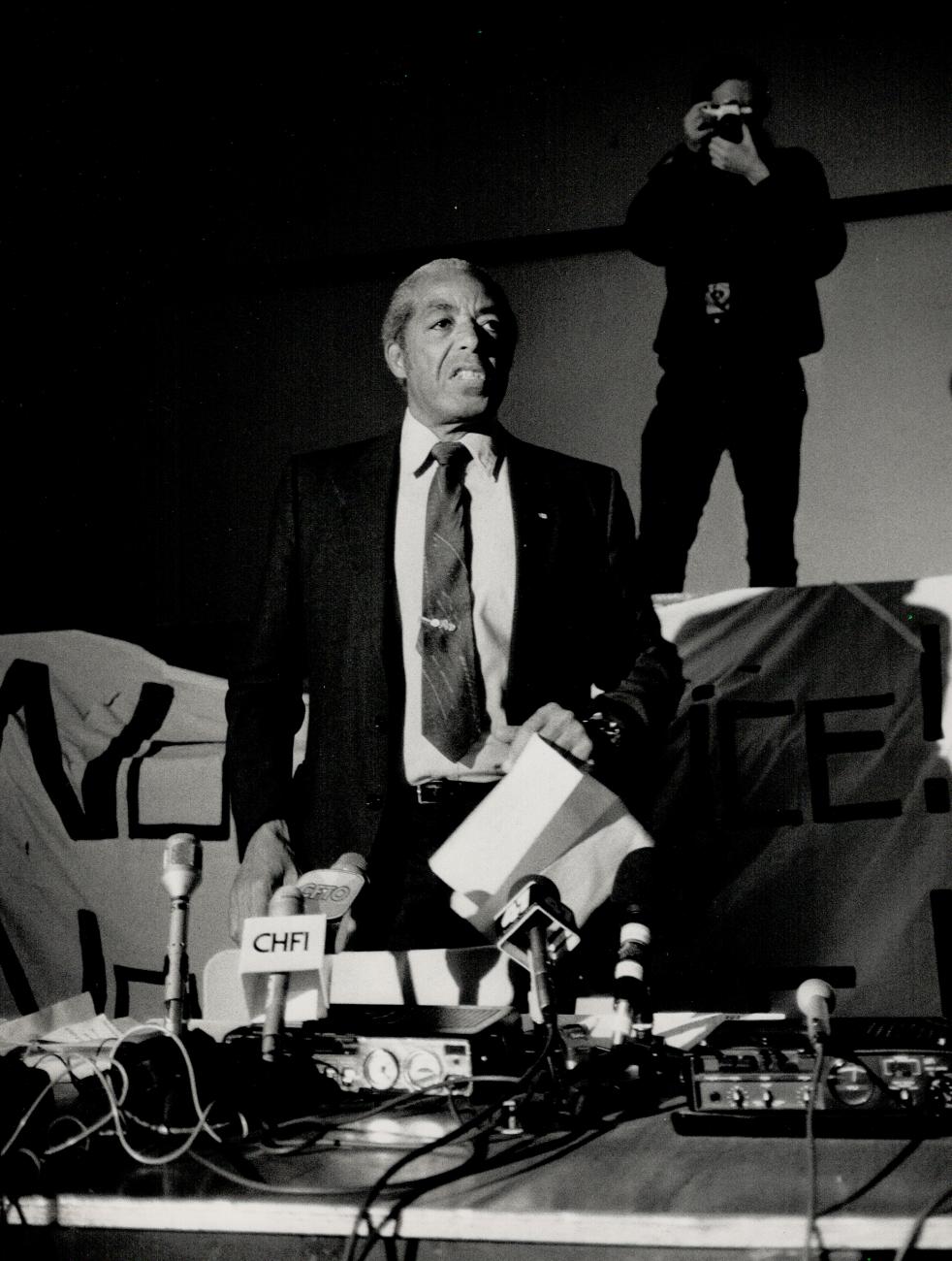Bromley Armstrong in a suit and tie holding some paper, standing with several microphones in front of him.