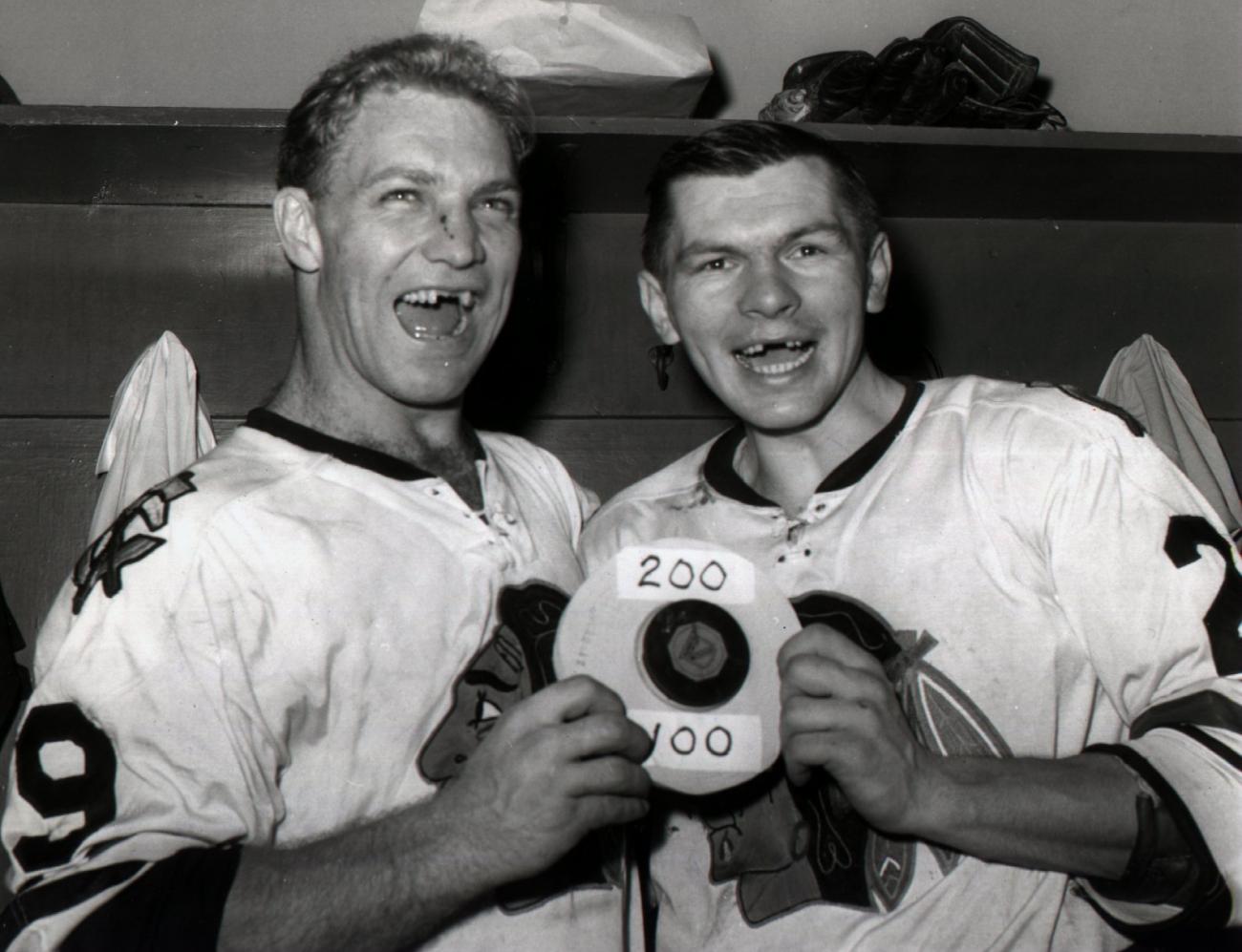 Stan Mikita poses smiling with another man in white team jerseys holding a puck, both with their upper front teeth missing.