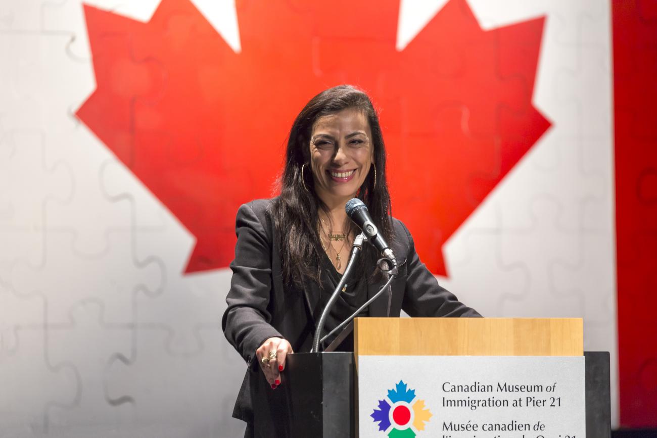 Carmen Aguirre at a podium in front of the Canadian flag.