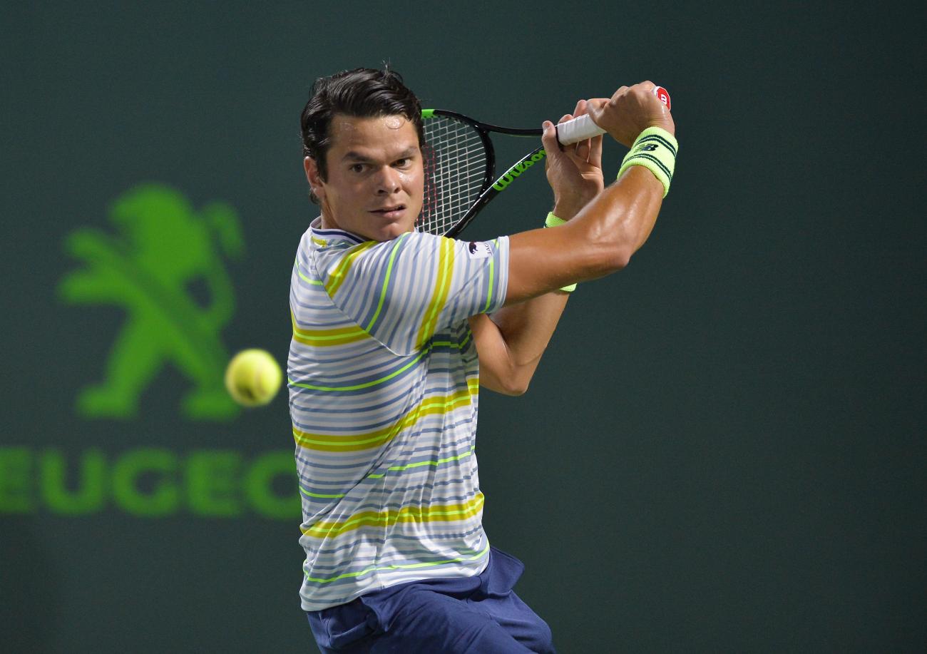 Milos Raonic holding a tennis racket swinging at a tennis ball in the foreground.