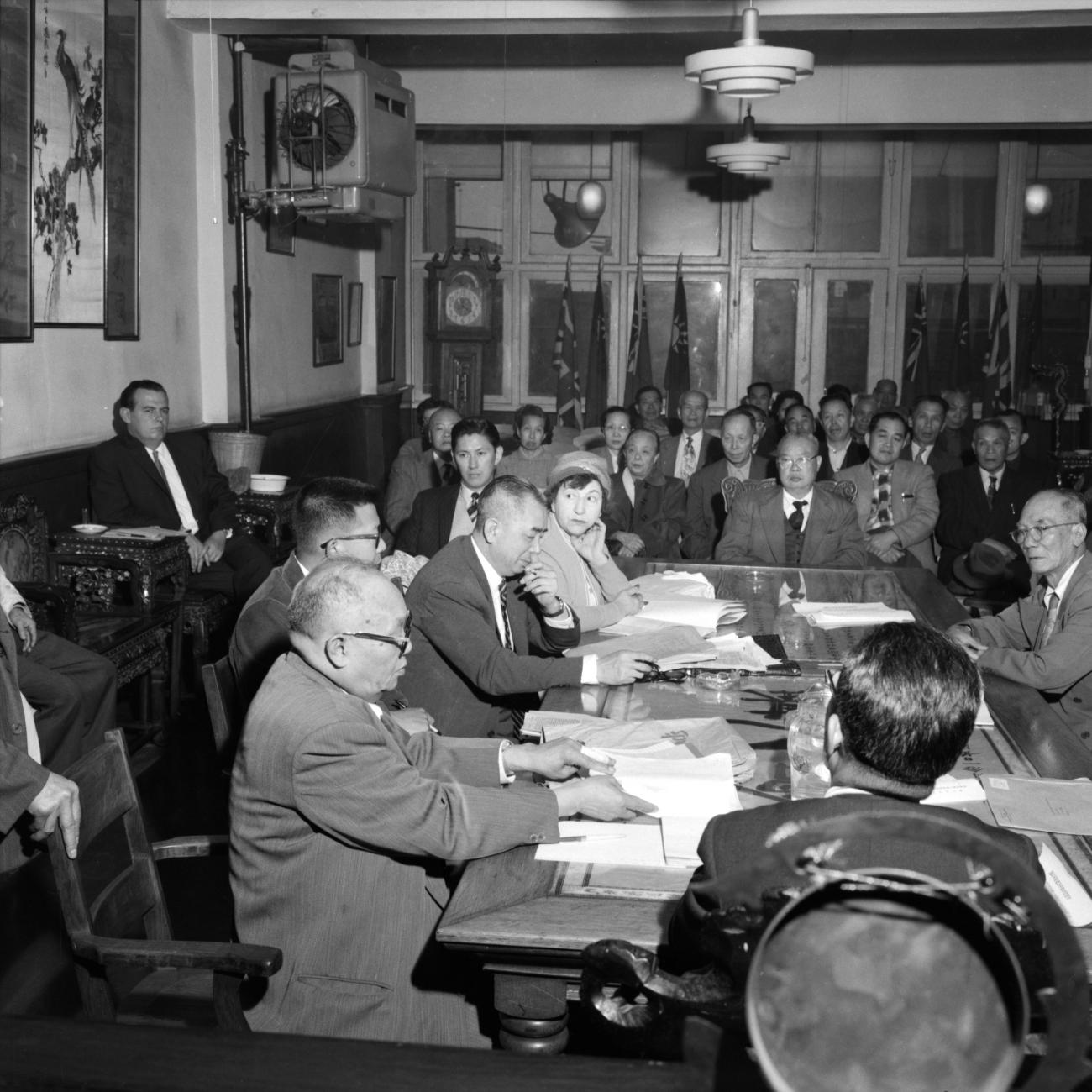 Wong Foon Sien sitting at a table with five other people with other people sitting on rows of chairs.