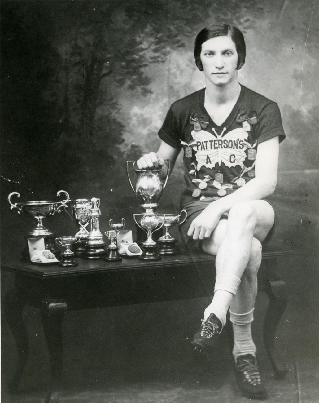 Bobbie Rosenfeld seated on a small table next to many trophies.