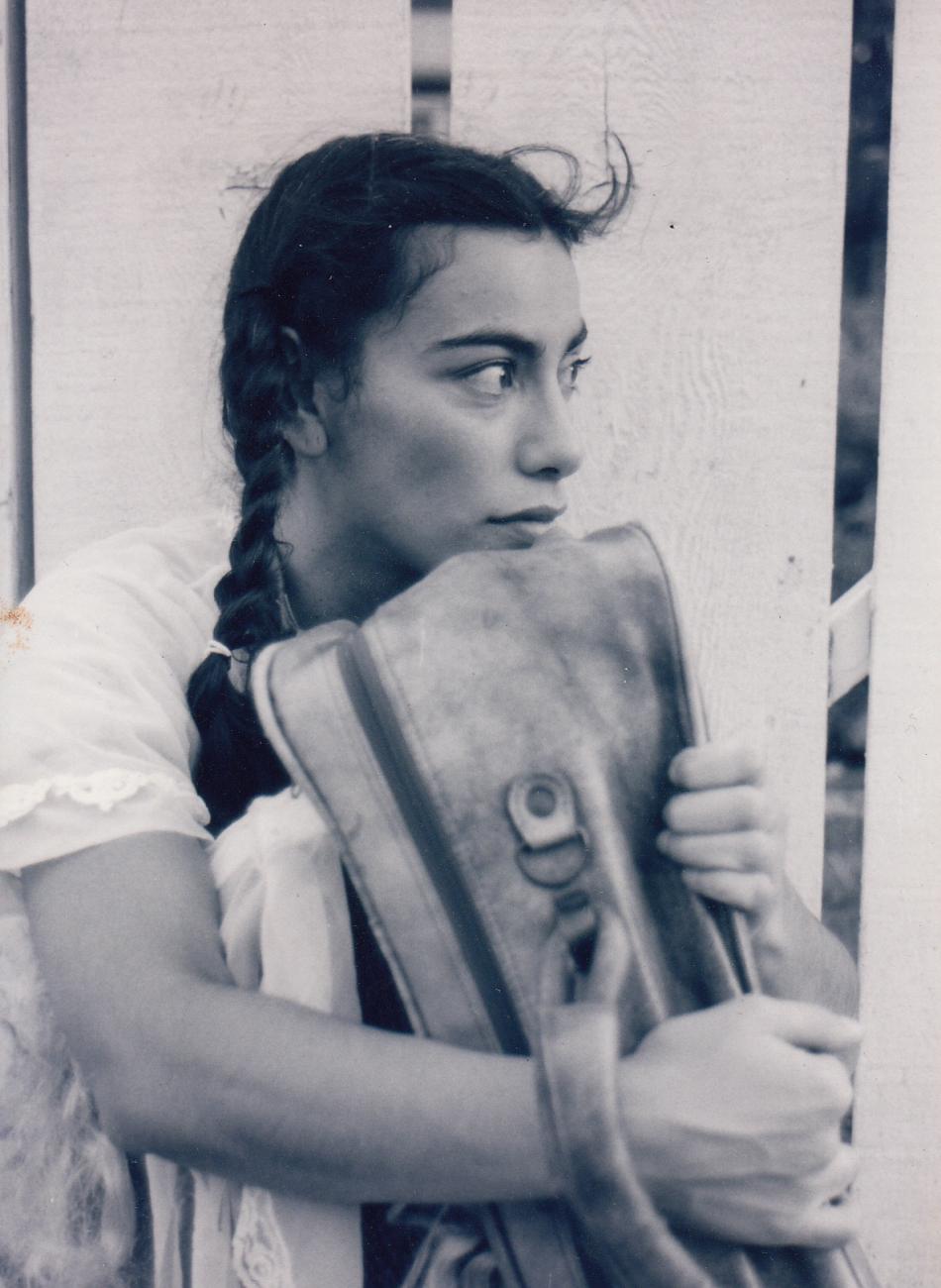 Carmen Aguirre in braids sitting hugging a suitcase.