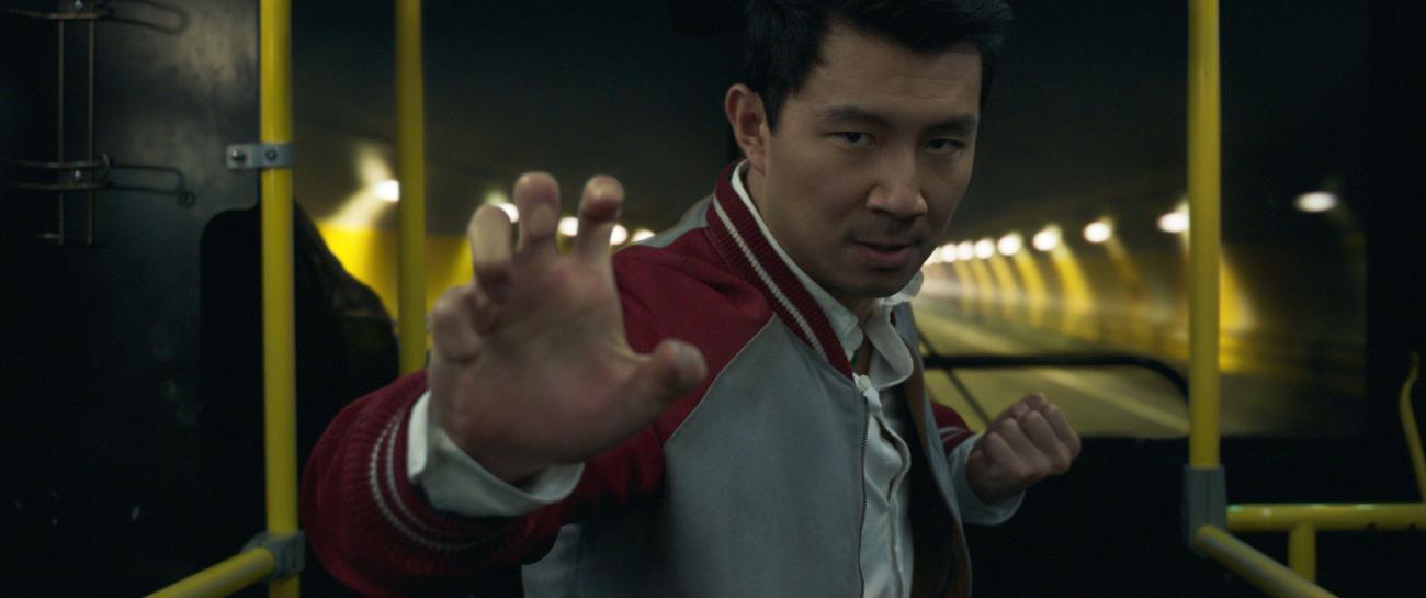 Simu Liu, as Shang-Chi, holds his right hand up, fingers spread, as though he is reading for a fight.