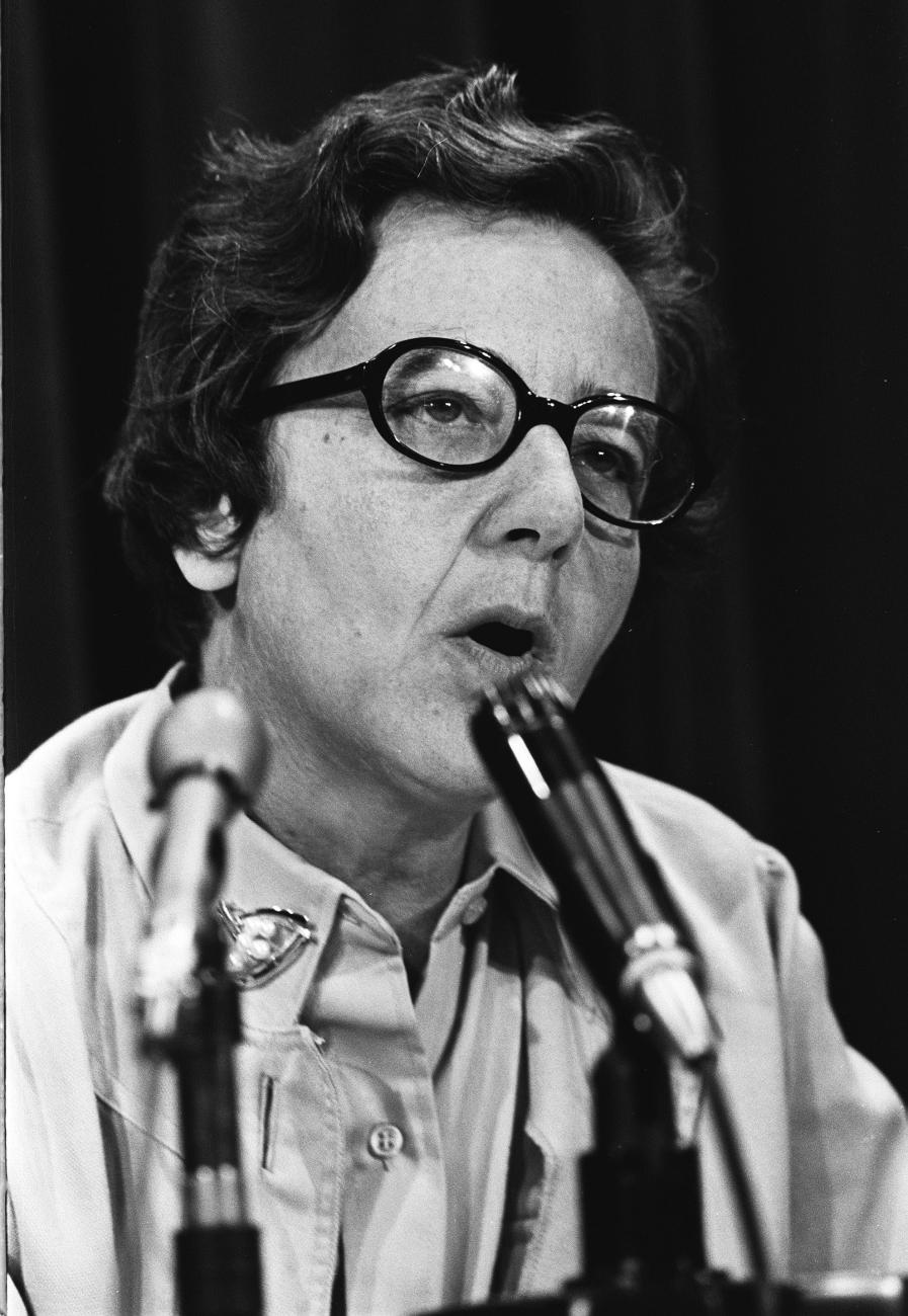 A woman speaks into microphones placed on the table in front of her