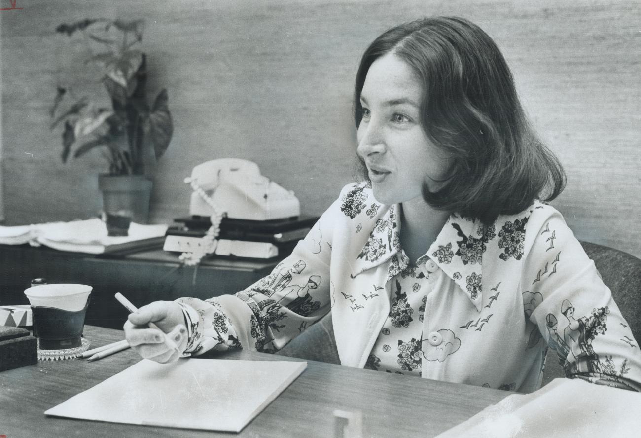 A woman sits at a desk with a pen in her hand and paper in front of her.