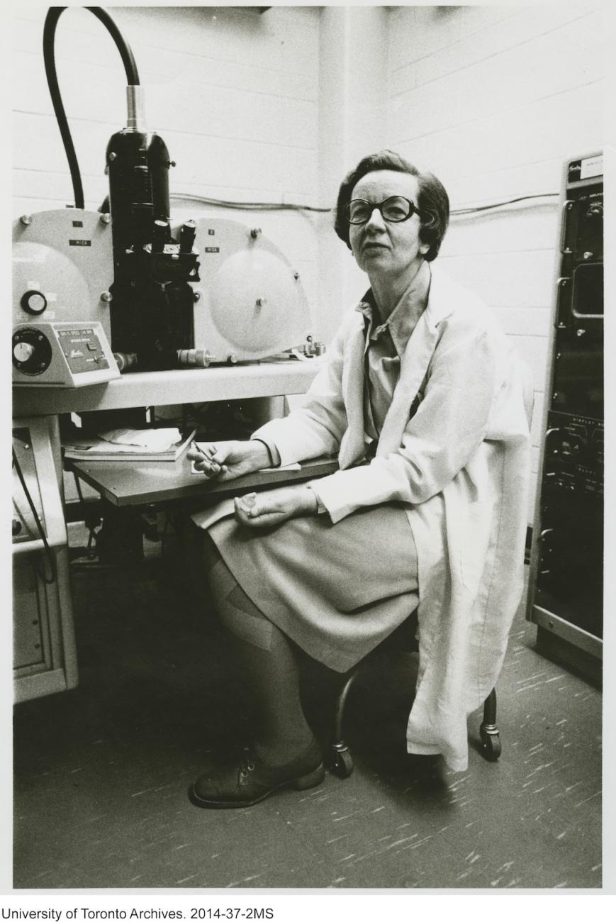 A black and white photo of a scientist in a lab coat, sitting in front of a large piece of lab equipment